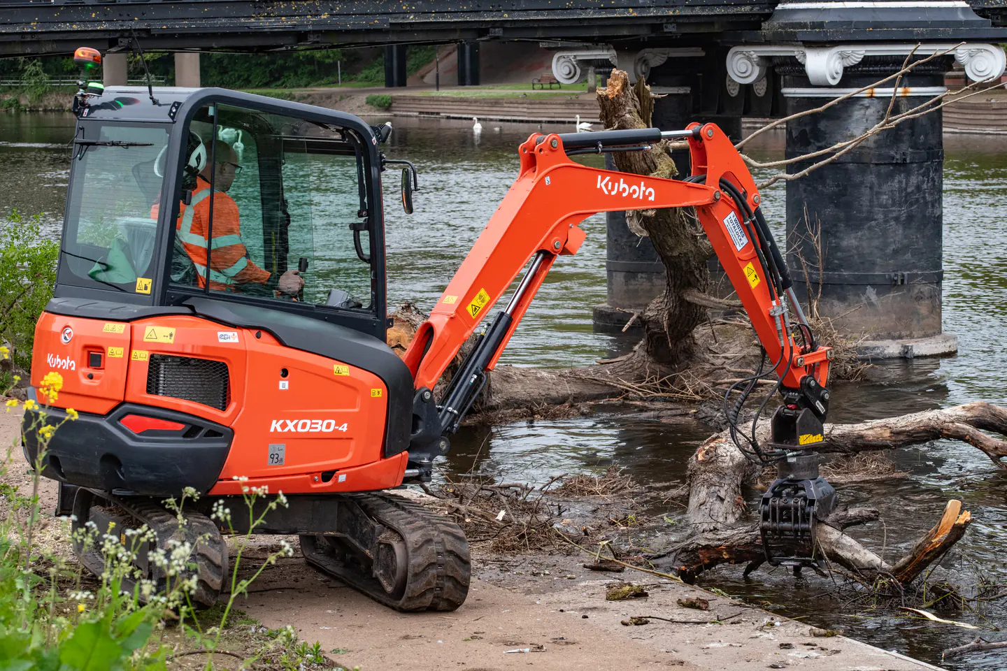 Excavator handling timber during tree work beside a bridge and waterway